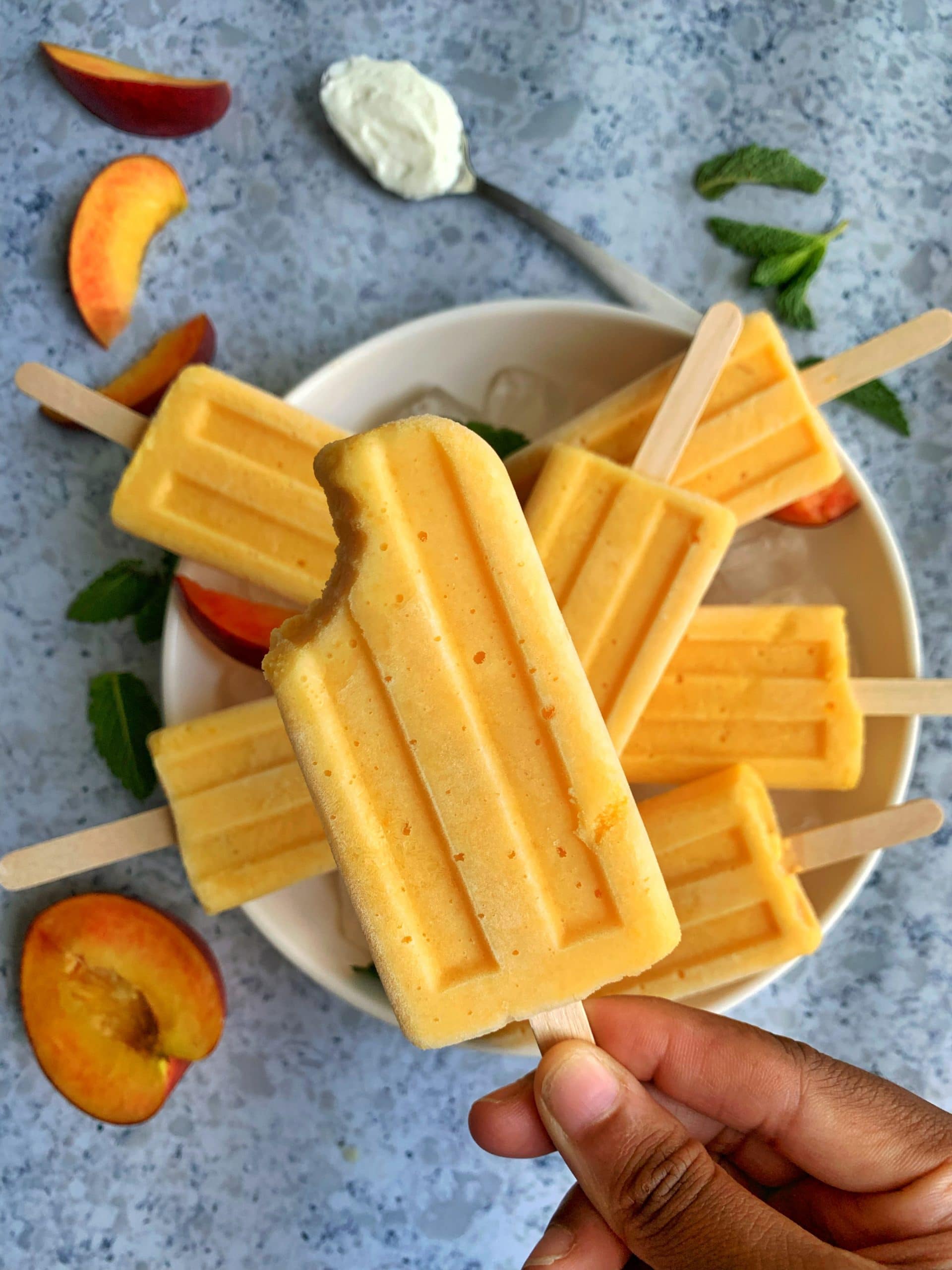 Homemade creamy peach popsicles on a bowl of ice with mint leaves, sliced peaches, and a spoon of greek yogurt in the background.