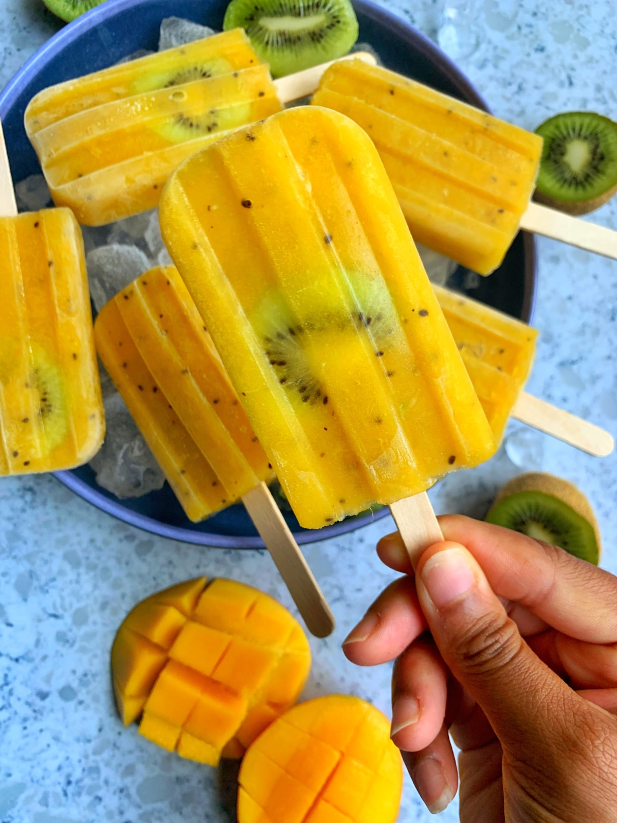 A hand holding a homemade mango kiwi popsicle with more treats in the background.