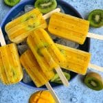 Homemade Mango-Kiwi Popsicles in a bowl of ice with extra mangoes and cut kiwi on the side.