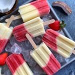 A tray of homemade Miami Vice Popsicles on a bed of ice with coconut halves and strawberries on the side.