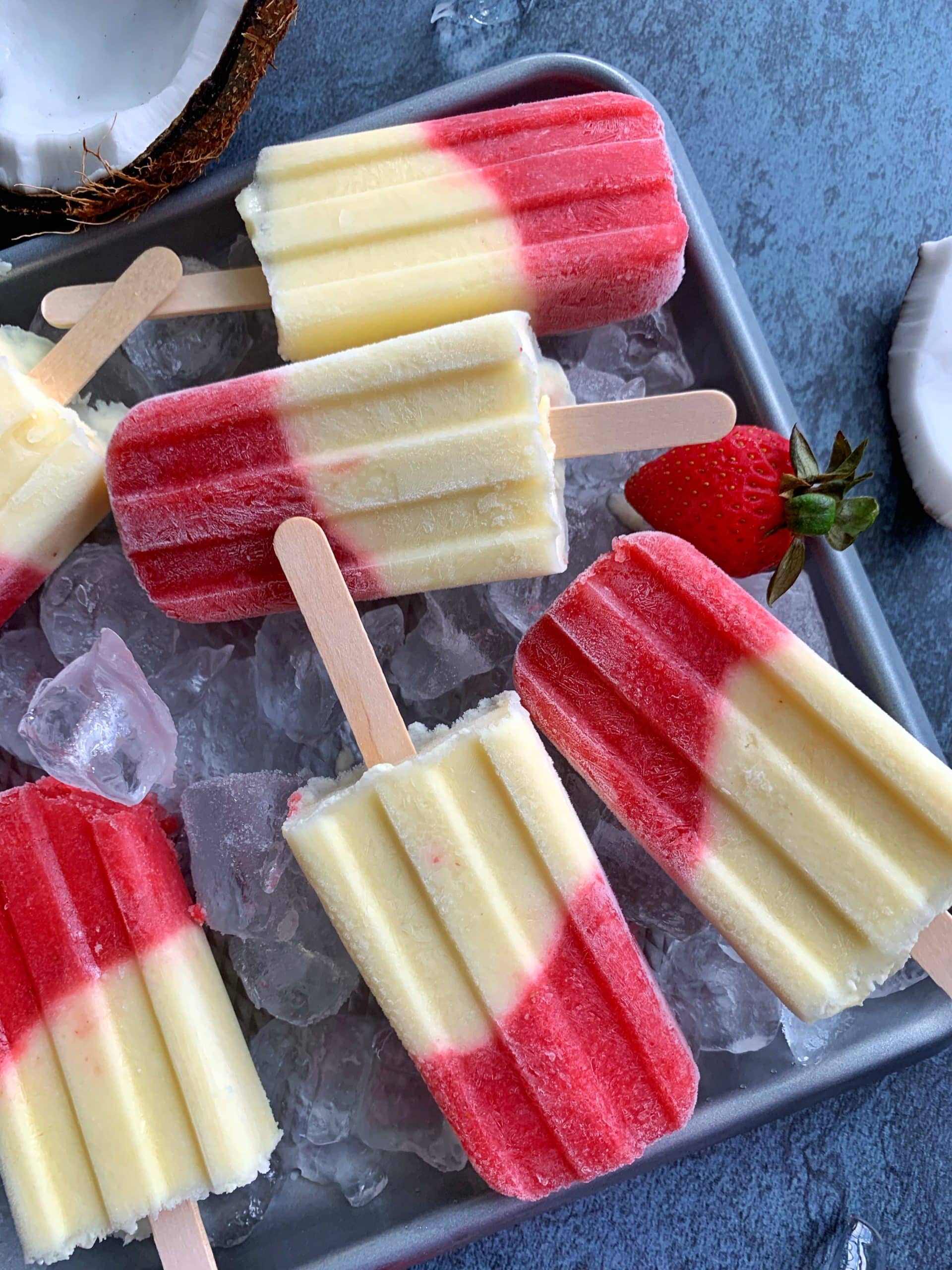 A tray of homemade Miami Vice Popsicles on a bed of ice with coconut halves and strawberries on the side.