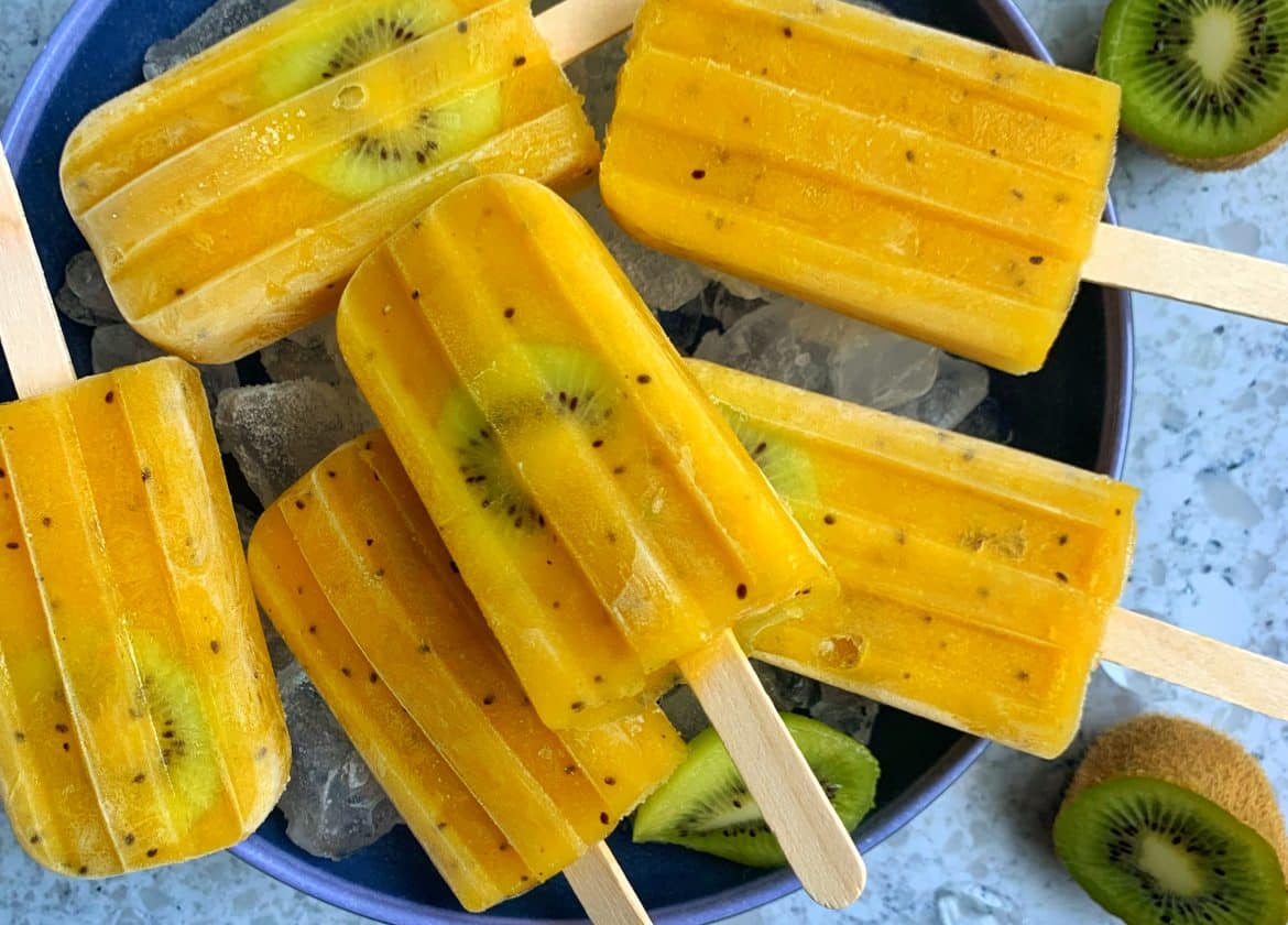 Homemade Mango-Kiwi Popsicles in a bowl of ice with extra mangoes and cut kiwi on the side.