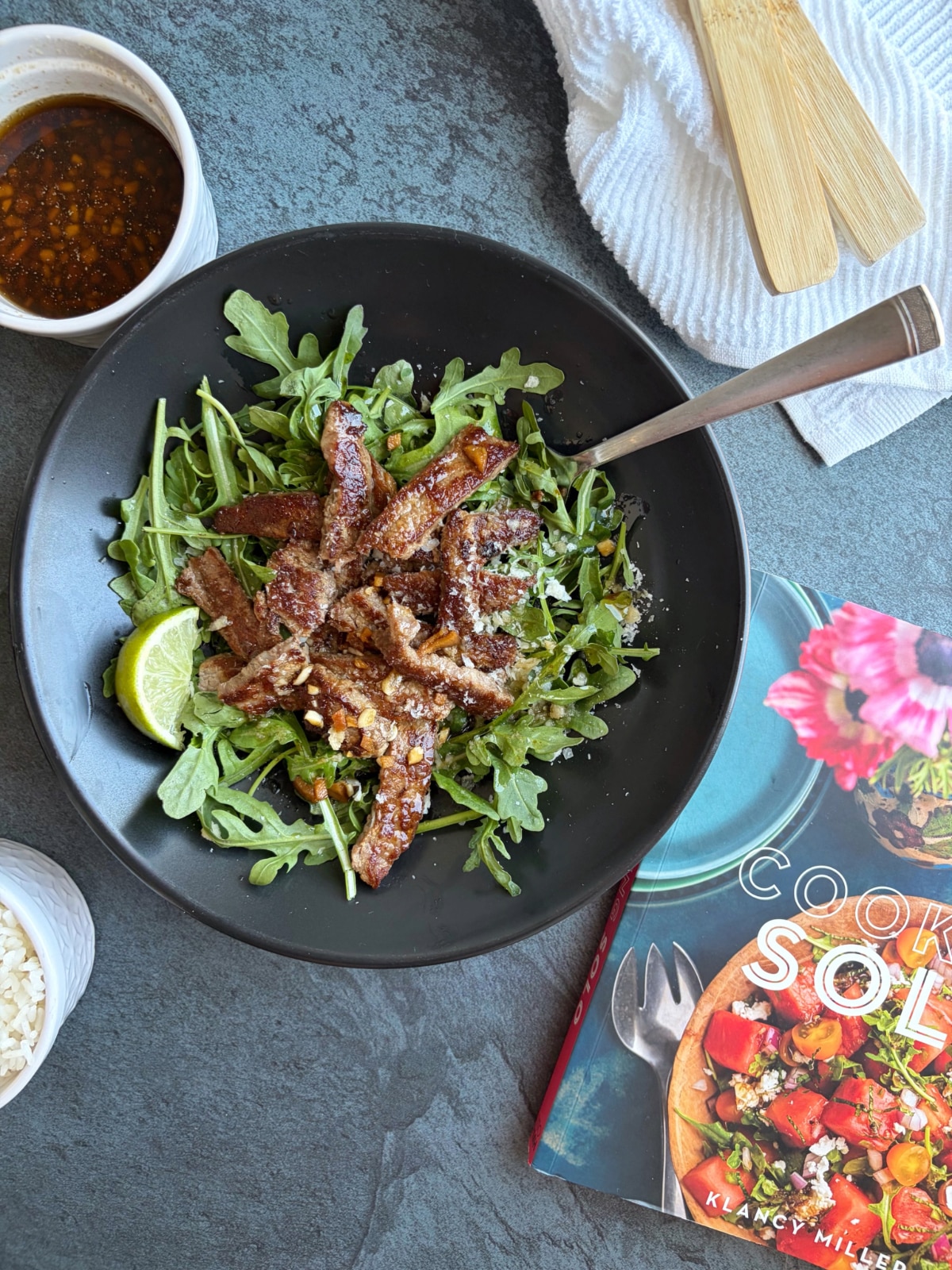 A bowl of beef stir fry over arugula with a cookbook next to it.