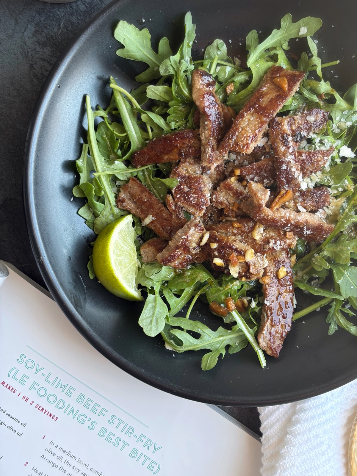 A bowl of beef stir fry over arugula, with a cookbook opened to the recipe next to it.
