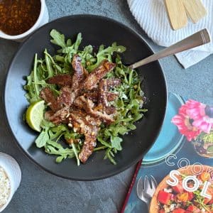 Seared steak strips on a bed of arugula with lime wedges on the side.
