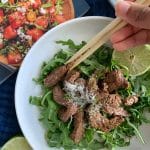 A hand picking up a piece of Soy Lime Beef Stir Fry with chopsticks. A cookbook in the background.