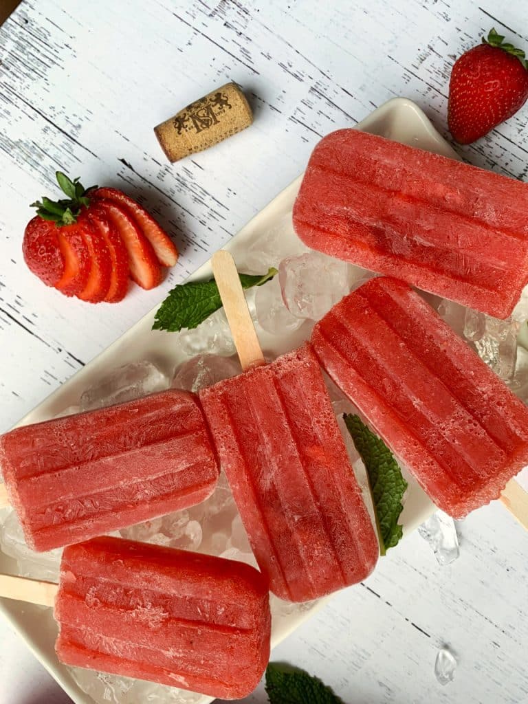 Homemade boozy Strawberry Rosé popsicles on a bed of ice with sliced strawberries on the side.