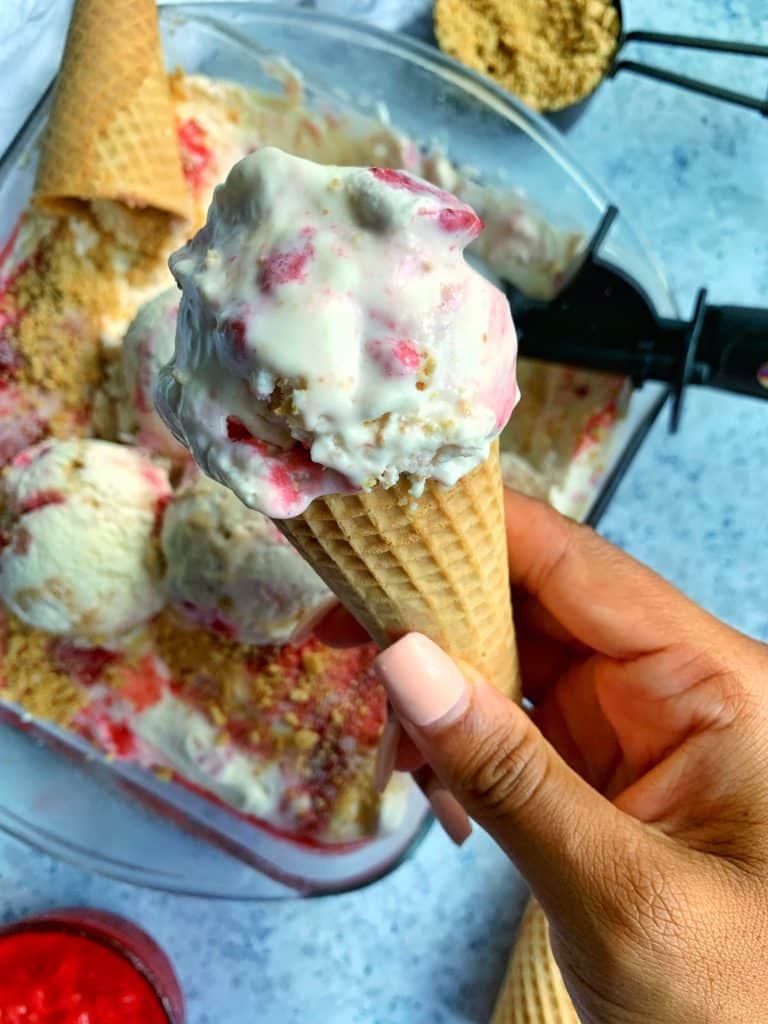 A cone of No-Churn Strawberry Cheesecake Ice Cream with more ice cream and strawberry compote in the background.