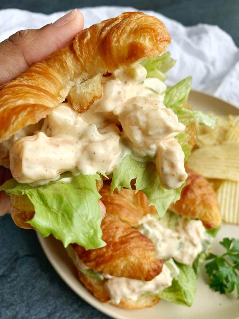 A hand holding a cold shrimp salad sandwich on a croissant with another sandwich and chips in the background.