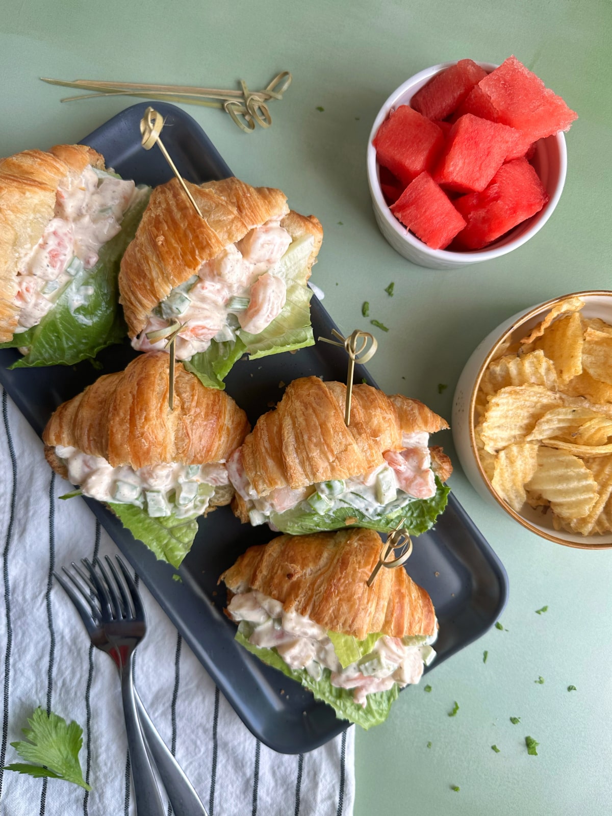A tray of shrimp salad stuffed inside mini croissants, with bowls of fresh watermelon and chips on the side.