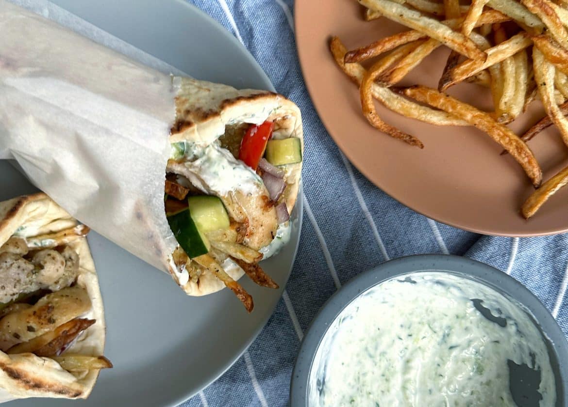 Two Greek Chicken Gyros wrapped in parchment paper with homemade fries, Greek salad, and homemade tzatziki sauce on the side.