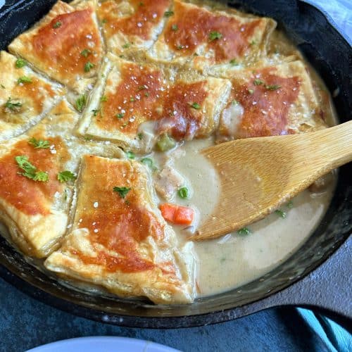 Chicken Pot Pie with Puff Pastry in a cast-iron skillet with a chunk sliced out and served on a nearby plate.