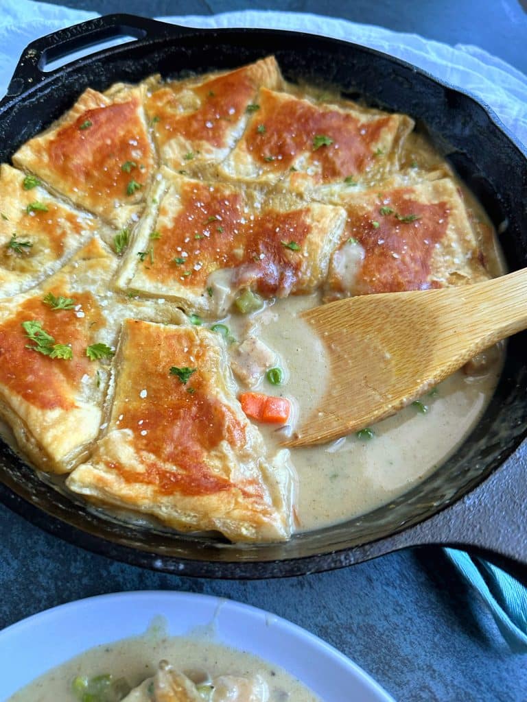 Chicken Pot Pie with Puff Pastry in a cast-iron skillet with a chunk sliced out and served on a nearby plate.