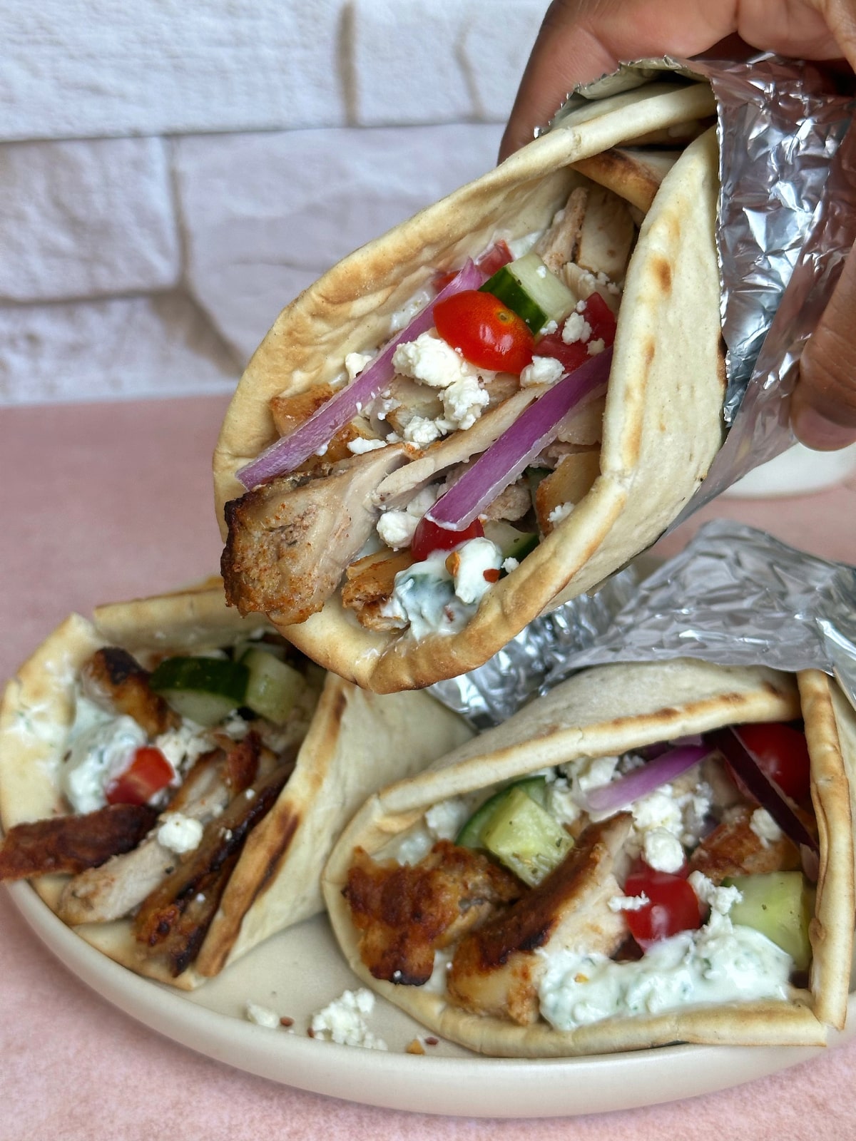 A hand picking up a foil-wrapped gyro wrap from a stack of them sitting on a plate.