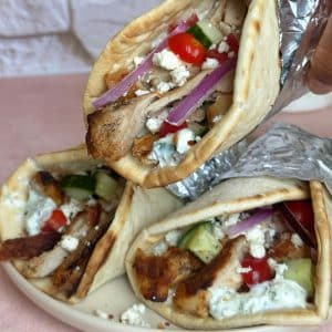 A plate of chicken gyros wrapped in foil, with a hand lifting one off of the others.
