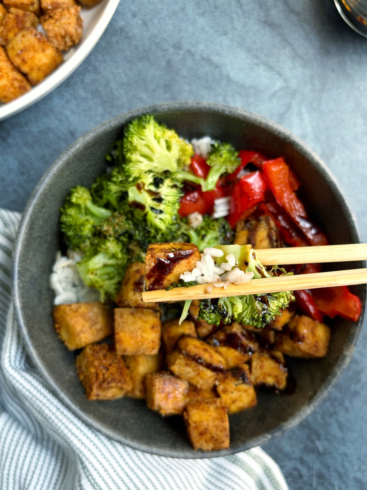 A pair of chopsticks holding up a bite of tofu, rice, and broccoli over the rest of the tofu power bowl.