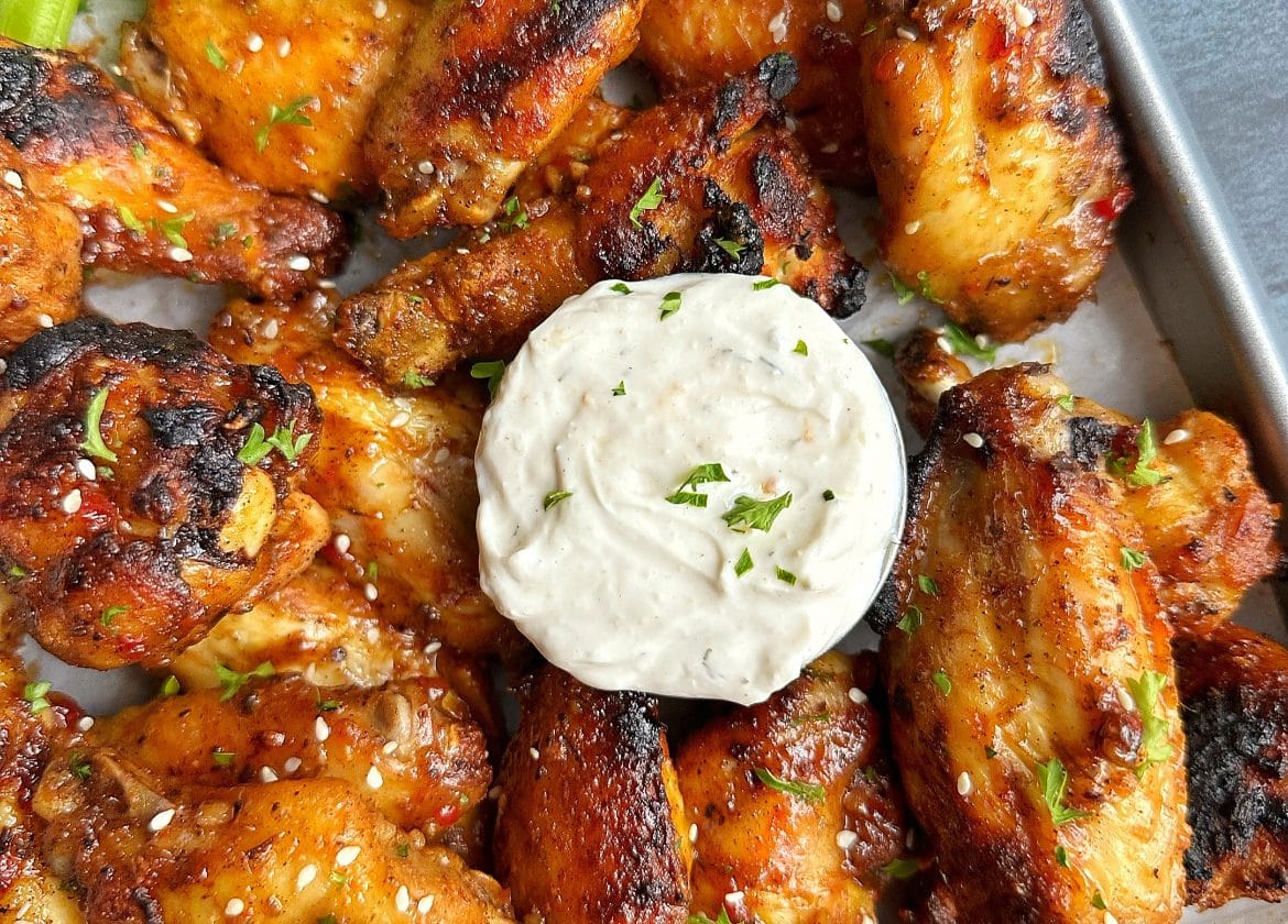 A tray of sweet chili jerk wings with a cup of ranch in the middle and celery sticks on the top left corner.