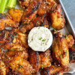 A tray of sweet chili jerk wings with a cup of ranch in the middle and celery sticks on the top left corner.