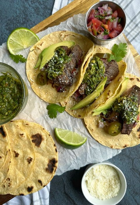 A tray of flank steak tacos with homemade chimichurri sauce on top, plus pico de Gallo and limes on the side.