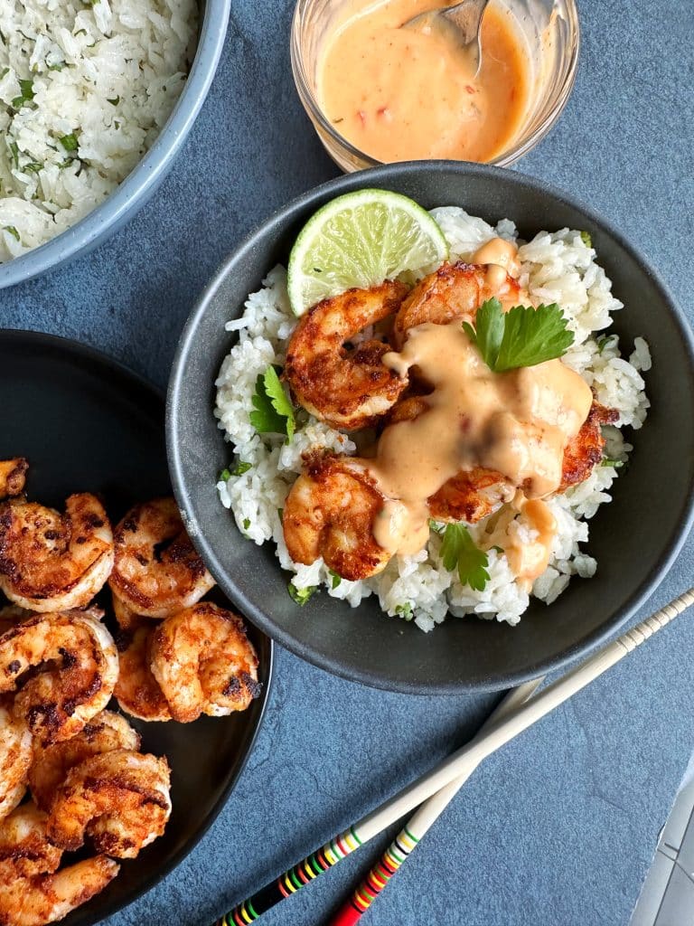 A bowl of Grilled Bang Bang Shrimp over rice with more shrimp, rice, and bang bang sauce on he side.