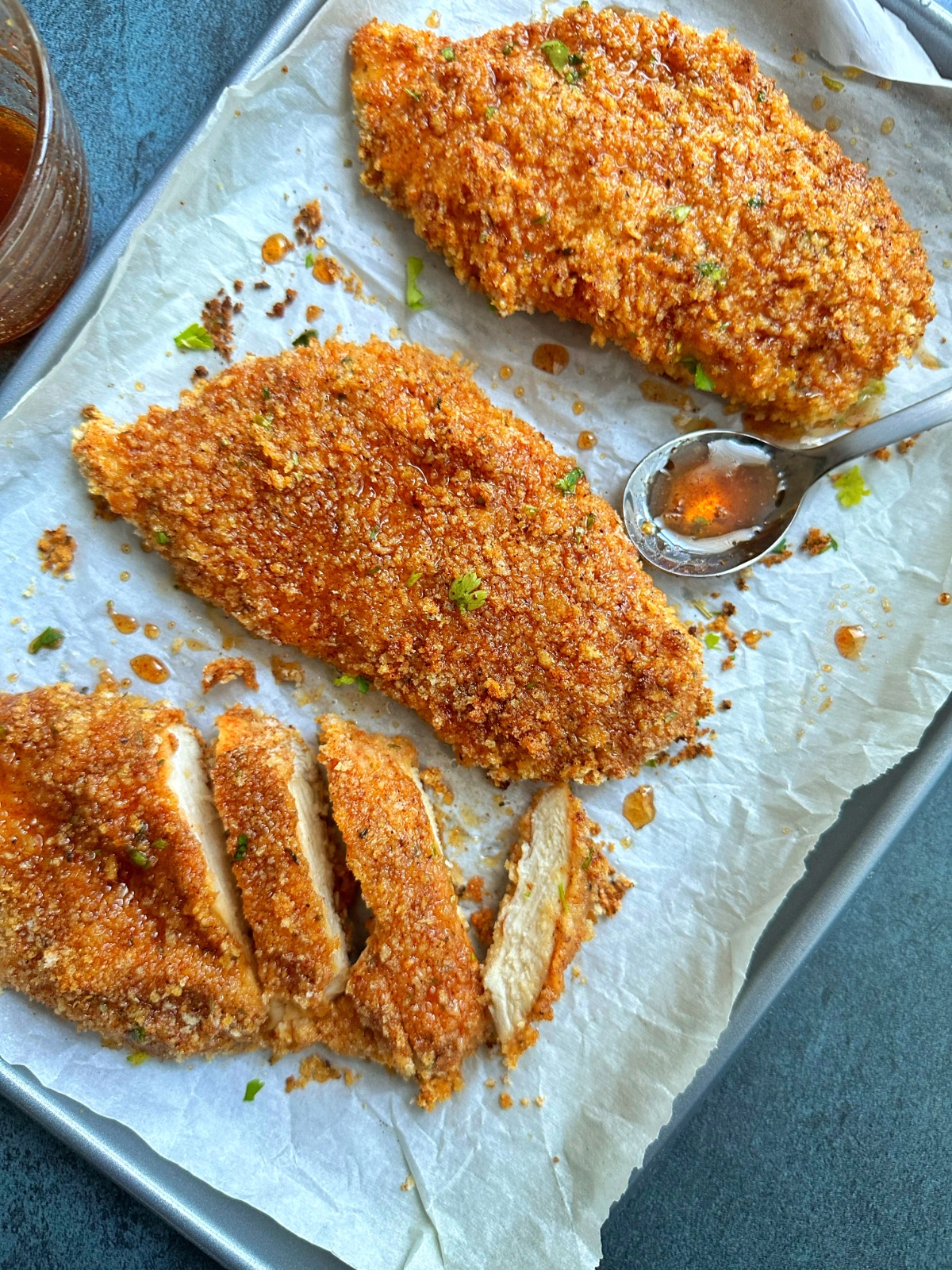 A parchment-lined pan of baked hot honey chicken with a spoon of hot honey on the side.