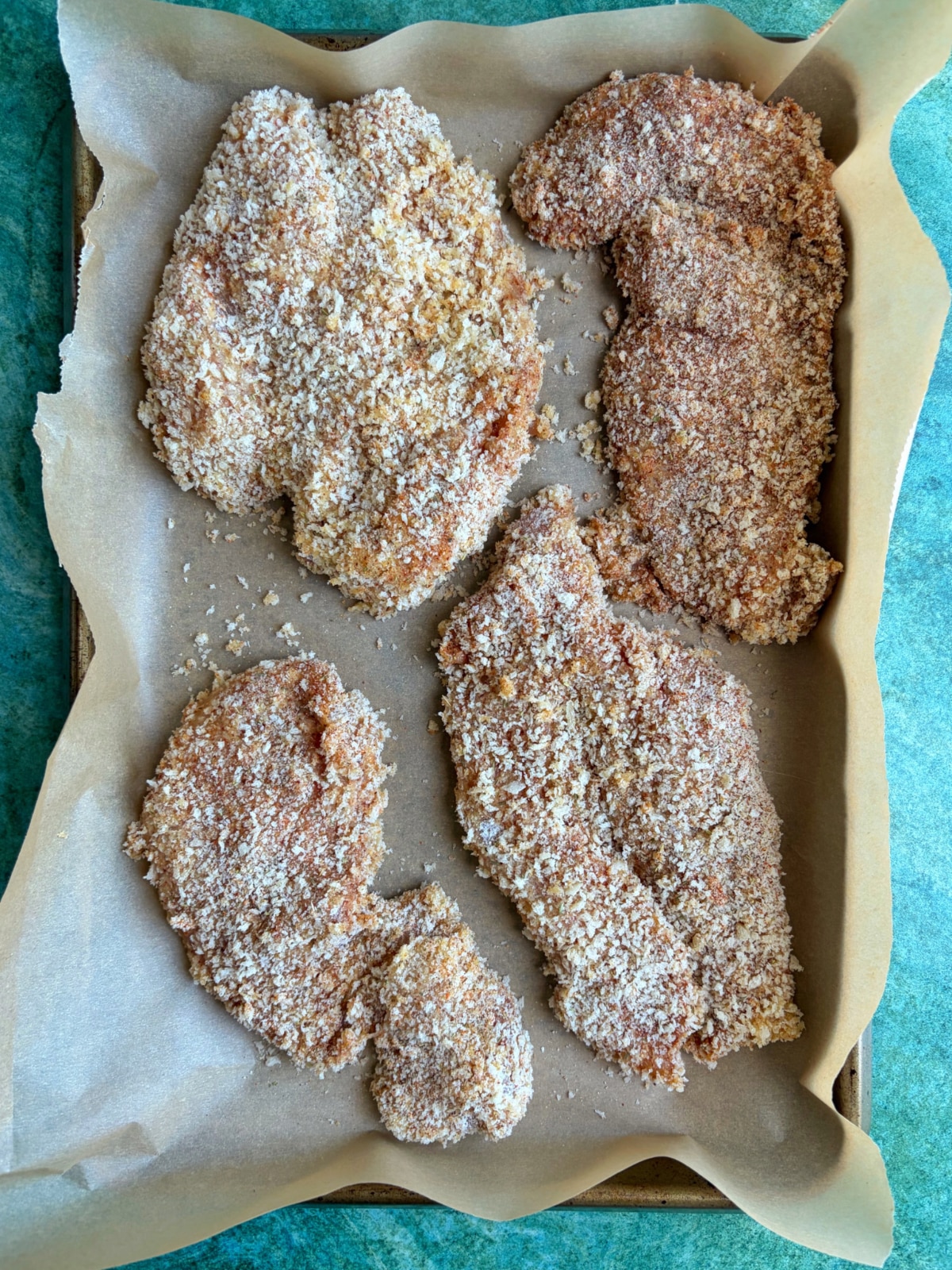 Chicken breasts breaded in panko breadcrumbs on a sheet pan before baking.