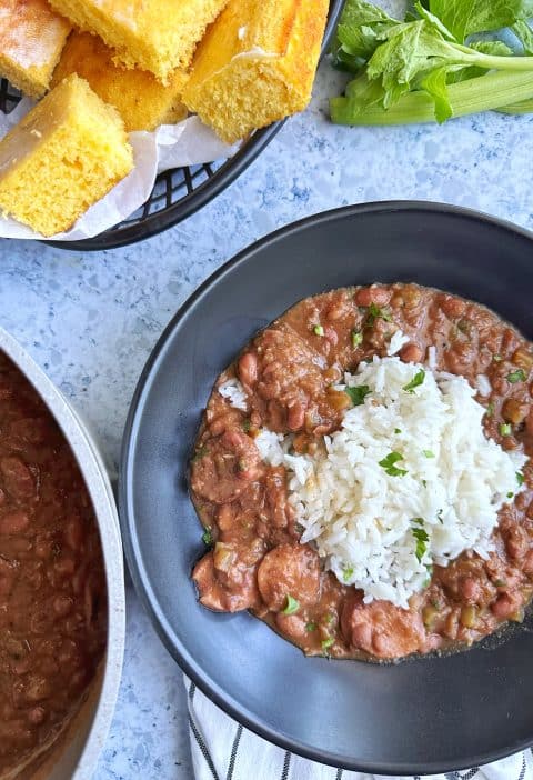 A black bowl filled with red beans & rice, with a basket of cornbread in the background.