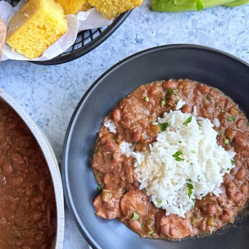A black bowl filled with red beans & rice, with a basket of cornbread in the background.