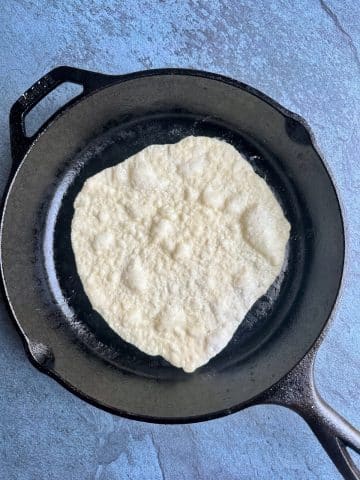 A cast iron skillet with a piece of naan cooking on top, with heat bubbles forming on the dough.