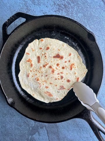 A piece of homemade naan being cooked on a cast iron skillet with a pair of tongs.
