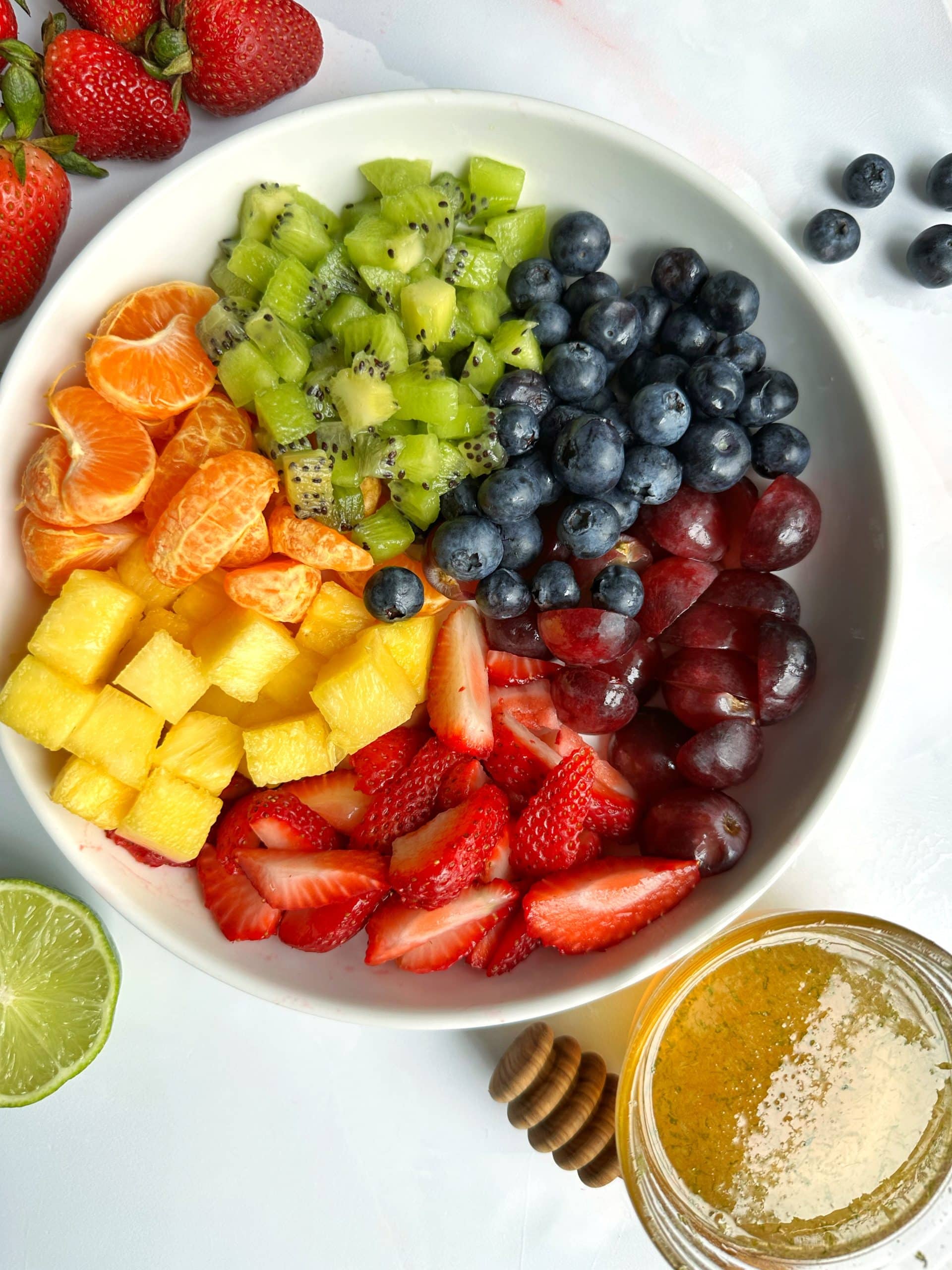 An overhead view of a bowl of rainbow fruit salad with a sauce bowl of agave-lime syrup on the side.