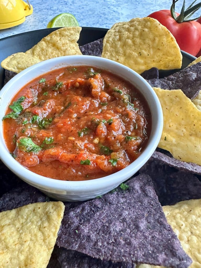 A bowl of restaurant-style, homemade fire roasted salsa with yellow & blue corn chips placed around it.