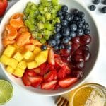 An overhead view of a bowl of rainbow fruit salad with a sauce bowl of agave-lime syrup on the side.