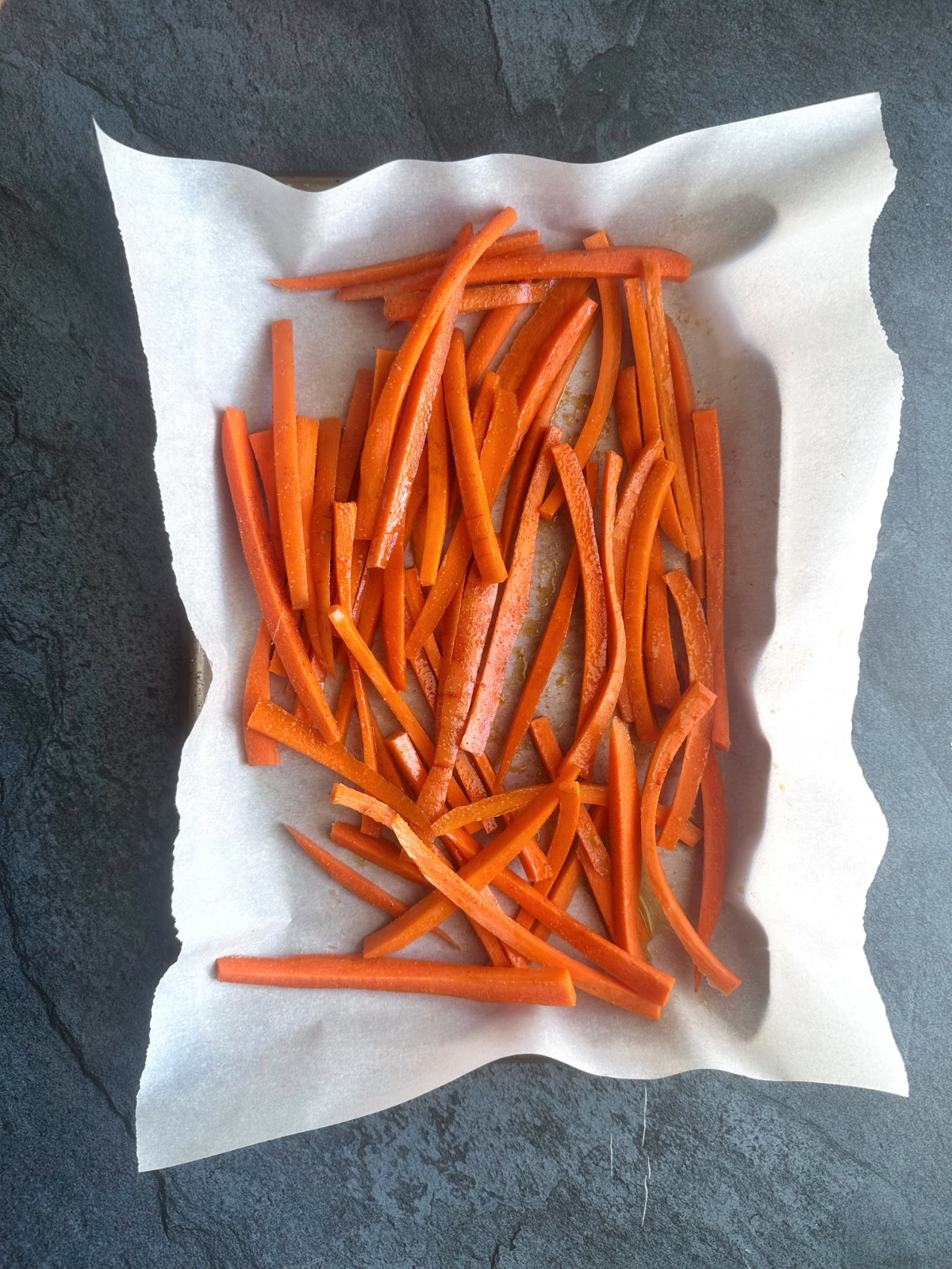 A parchment-lined pan of unbaked carrots cut into matchstick shapes.