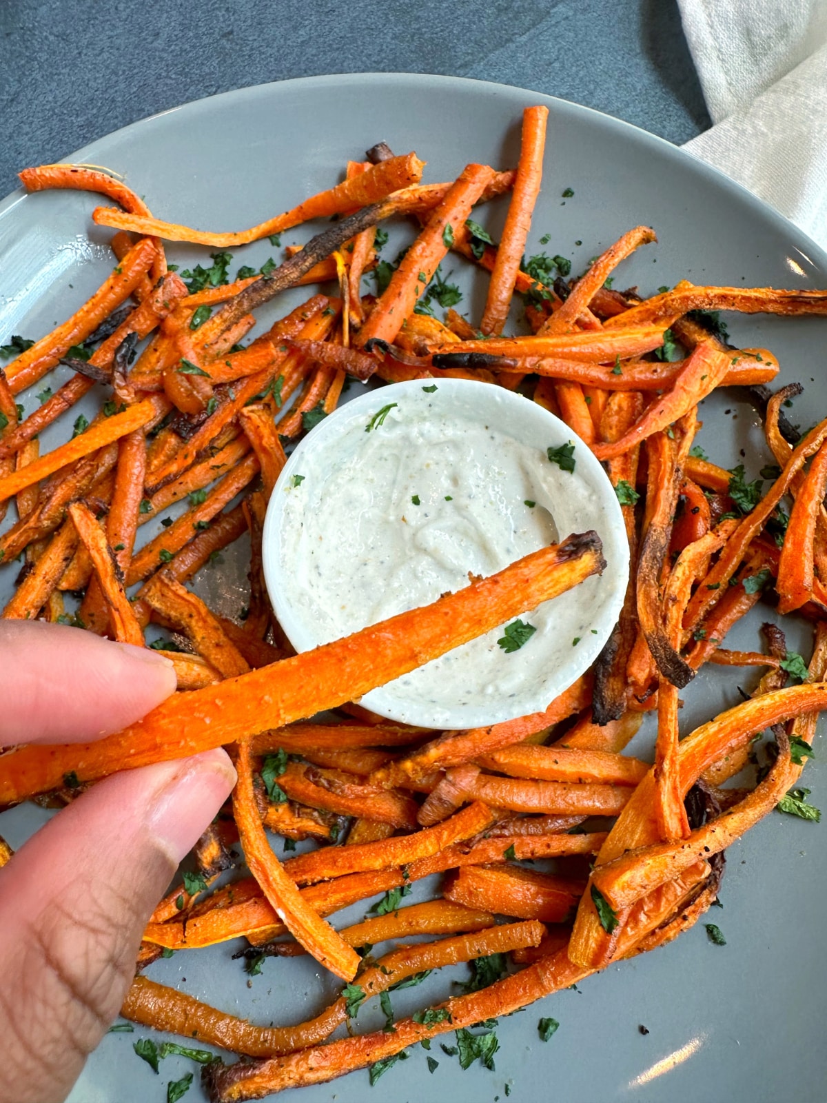 A hand holding a crispy baked carrot fry with a bowl of crispy baked carrot fries served with a white dipping sauce in the background.