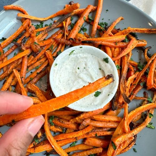 A hand holding a crispy baked carrot fry with a bowl of crispy baked carrot fries served with a white dipping sauce in the background.