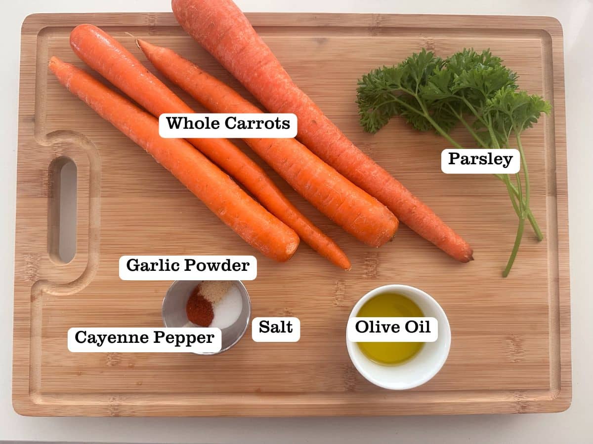 A wooden board containing whole carrots, parsley, olive oil, and seasonings in a small bowl.