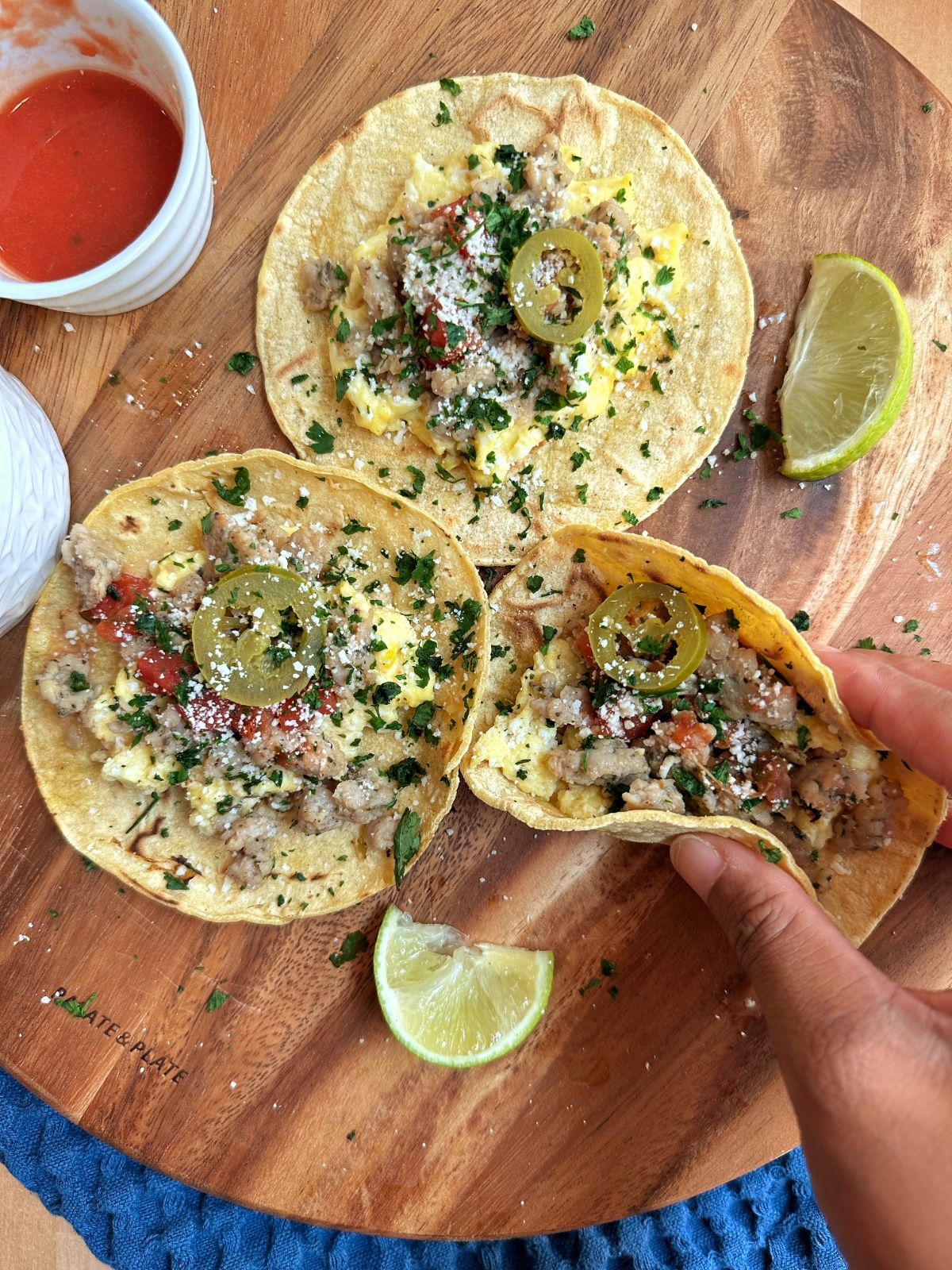 A wooden board containing three breakfast tacos with one being picked up by a hand.