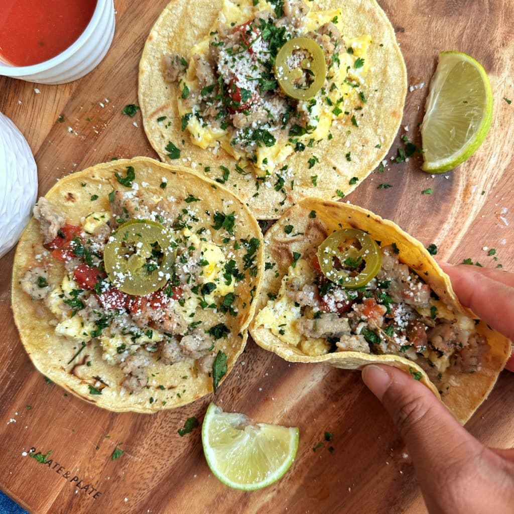 A platter with 3 homemade breakfast tacos and a hand picking one of them up.