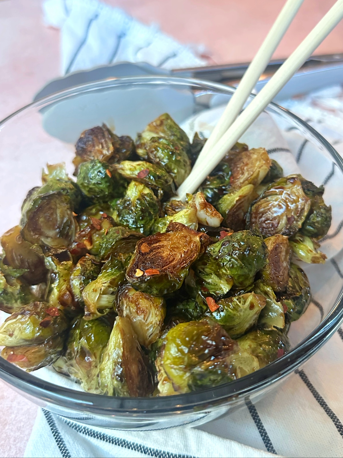 A clear bowl of roasted balsamic brussels sprouts with chopsticks sticking out of it.