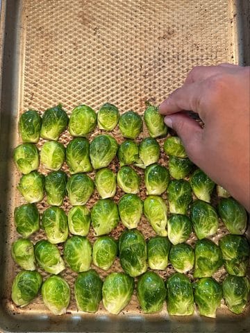 A hand placing halved brussels sprouts, cut side down, onto a sheet pan.