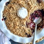 A white circular pan with Triple Berry Crumble inside, an ice cream scoop on top, and a serving spoon sitting in a corner.