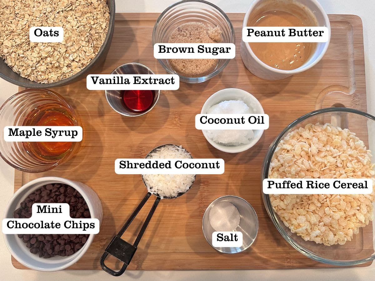 A wooden board filled with ingredients for homemade granola bars, including bowls of cereal, maple syrup, coconut oil, and brown sugar.