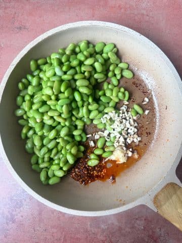 A skillet with defrosted shelled edamame, garlic, and chili sauce.