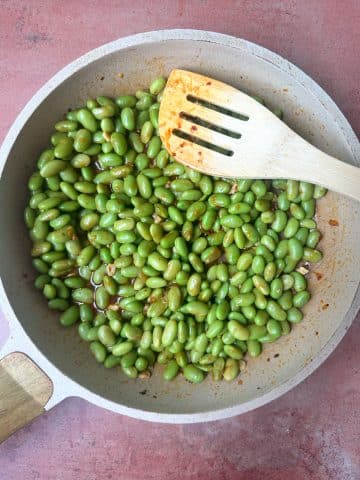 A skillet filled with chili crisp edamame and a wooden slotted spatula.