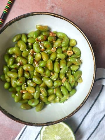 A bowl filled with chili lime edamame with a slice of lime and chopsticks on the side.