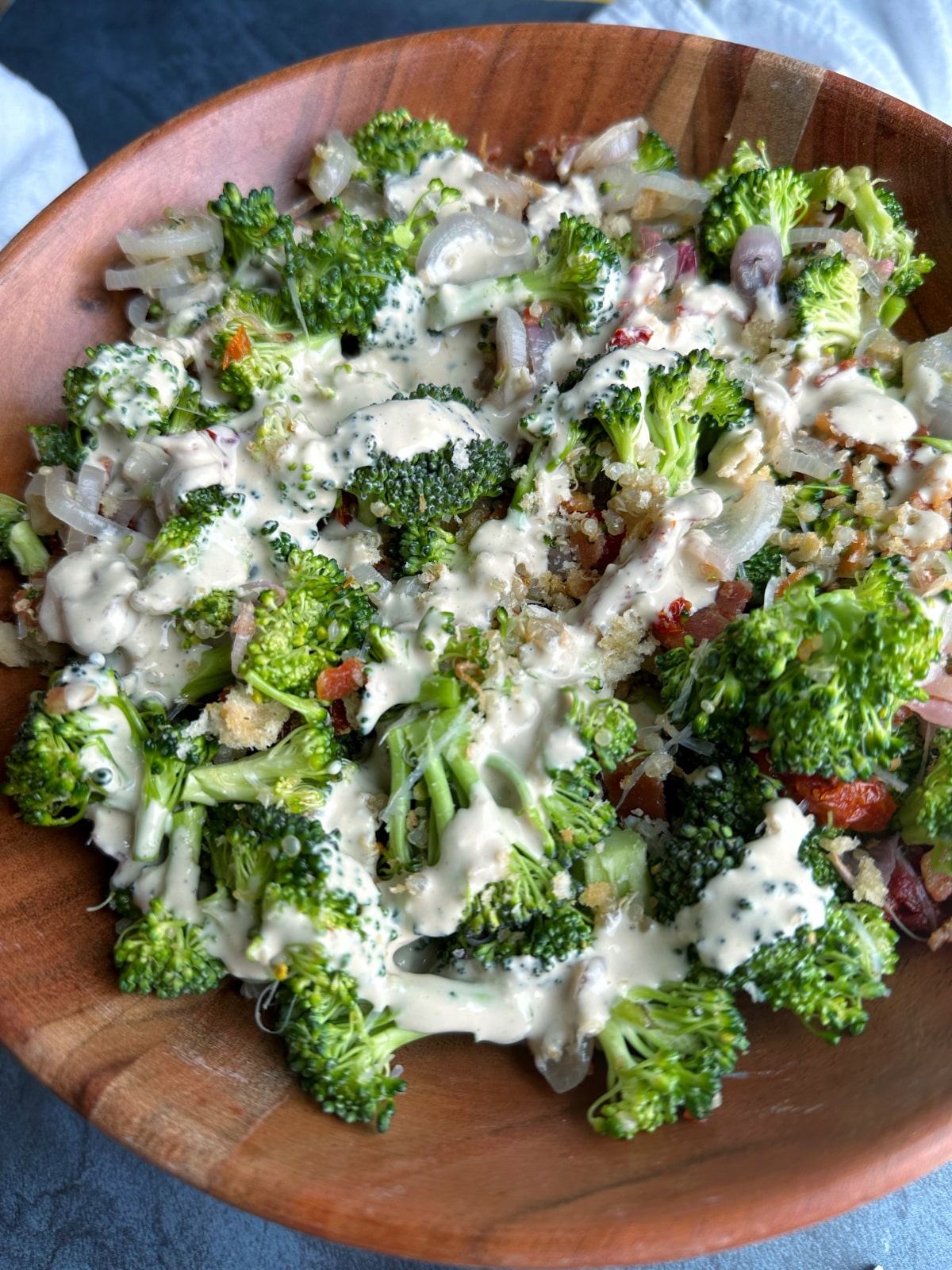 A wooden bowl of Broccoli Salad with bacon, sun-dried tomatoes, quinoa, croutons, shallots, and caesar dressing drizzle.