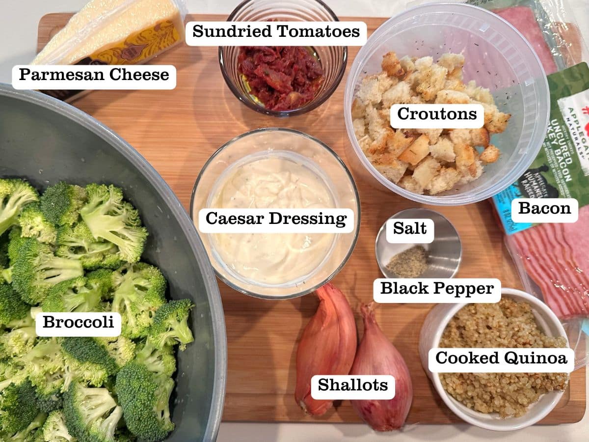 A wooden board containing ingredients for a salad, including caesar dressing, broccoli, croutons, and quinoa.
