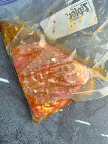 A flank steak covered in liquid marinade in a plastic storage bag.