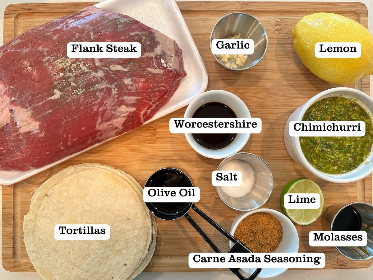A board containing bowls of ingredients for steak tacos, including chimichurri, flank steak, a lemon, and molasses.